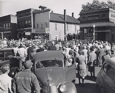 A crowd gathered in the street and in front of storefronts watching a speaker in the distance.