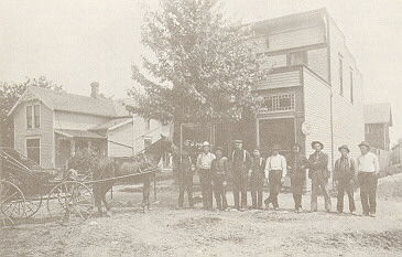 A row of men and a horsedrawn carriage stand in front of a pair of buildings.