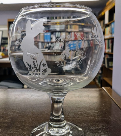 A clear glass goblet with an etched design of a rabbit and flowers on its surface, placed on a wooden table in front of bookshelves.