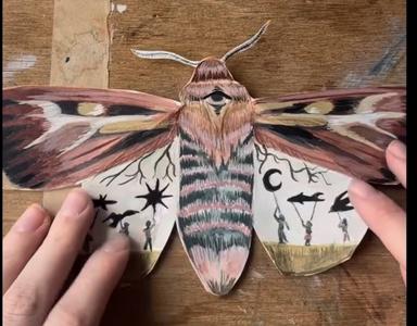 Two hands unfurl the wings of a crafted watercolor moth, revealing the images hidden beneath.