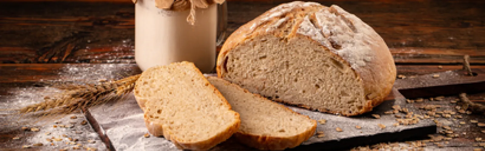 A cut loaf of sourdough bread atop a cutting board, next to a jar of sourdough starter.
